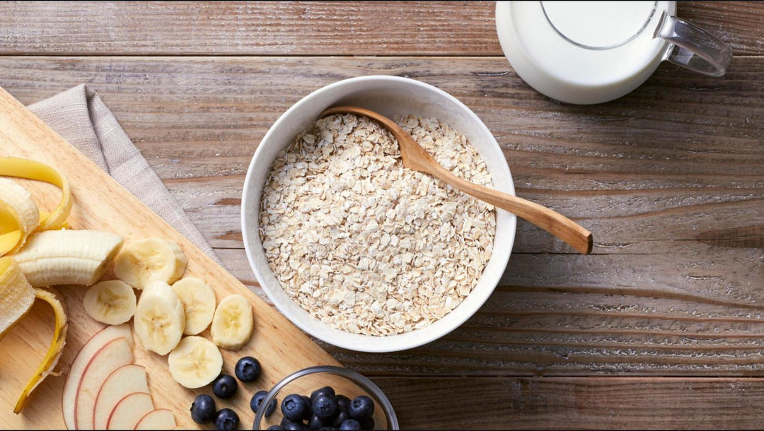 A bowl of oats with a wooden spoon beside sliced banana, apple and fresh blueberries and a pitcher of milk on a warm wooden kitchen table, a simple balanced breakfast for early recovery